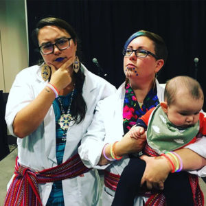 Co-hosts of the podcast Métis in Space, dressed in labcoats, Métis sashes and other futuristic accoutrements, with little space Métis baby in arms.